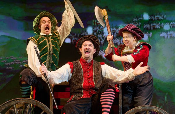 Actors in tudor costume are mid-attack, raising a sword and an axe respecitively above a third actor, sat on a cart holding a scepter, arms spread wide, mouth open in song.