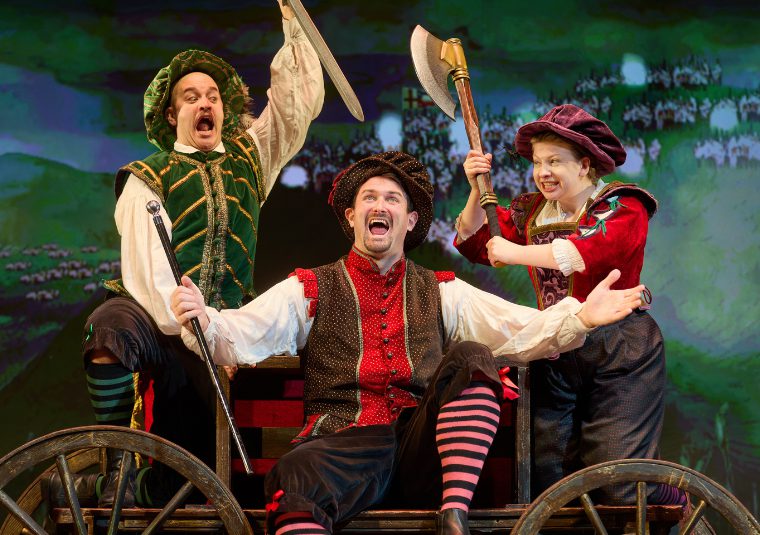 Actors in tudor costume are mid-attack, raising a sword and an axe respecitively above a third actor, sat on a cart holding a scepter, arms spread wide, mouth open in song.