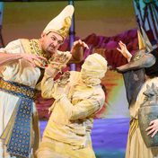 A Pharaoh leans over menacingly towards a mummy while an actor dressed as the god Anubis with a dog head and lose flowing costume holding a canopic jar.