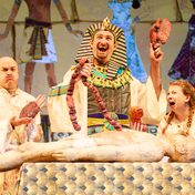 A pharaoh smiles holding up an organ from a mummy lying in front of him as he removes other body parts. Two servants stand on either side of him cry and looking like they are about to vomit.