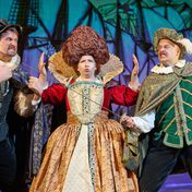 Two actors in tudor costume fight across a third actor who is dressed as a queen with big hair sewn into shape. The fighters are poised, faces aggressive, one raises their fists and the other points a rapier, the queen keeps them apart, hands out, lips pursed.