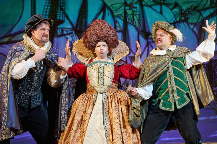 Two actors in tudor costume fight across a third actor who is dressed as a queen with big hair sewn into shape. The fighters are poised, faces aggressive, one raises their fists and the other points a rapier, the queen keeps them apart, hands out, lips pursed.