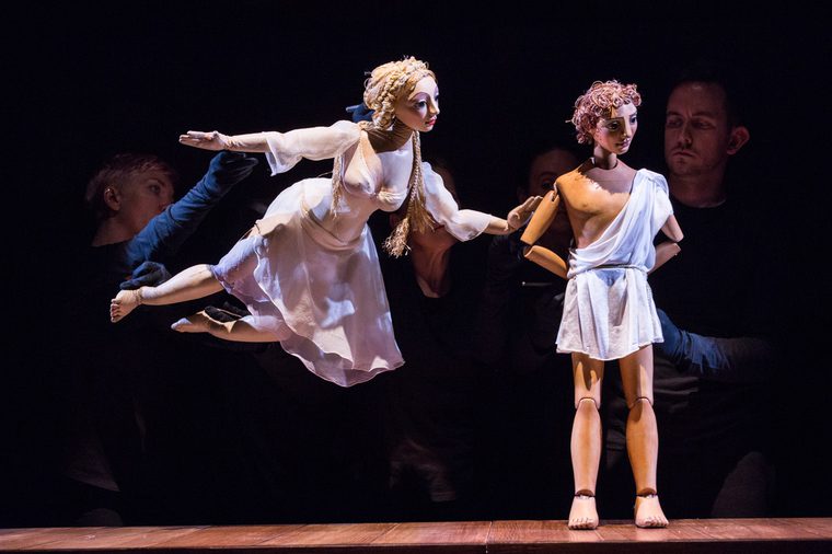 Venus flies to touch Adonis's arm where he's stood in contemplation. They are wood and fabric puppets, puppeteers barely visible in black gloves.
