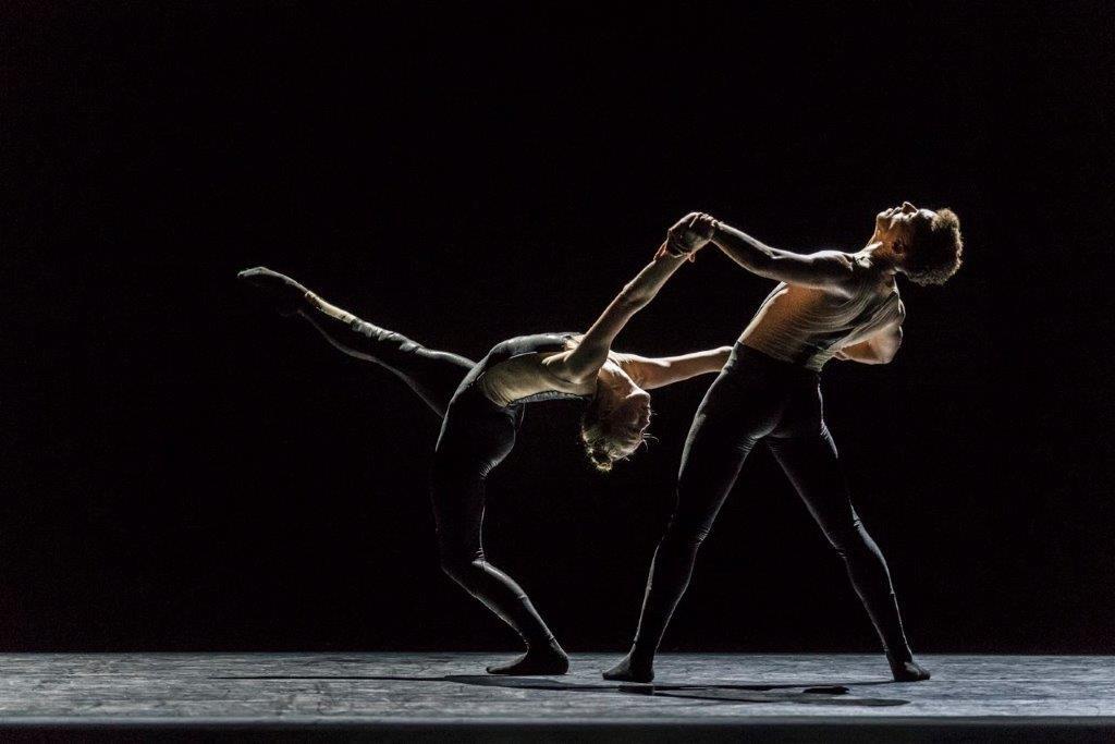 Two ballet dancers on stage, one leaning backwards with leg kicked up in the air while being supported by a dancer behind holding their hands.