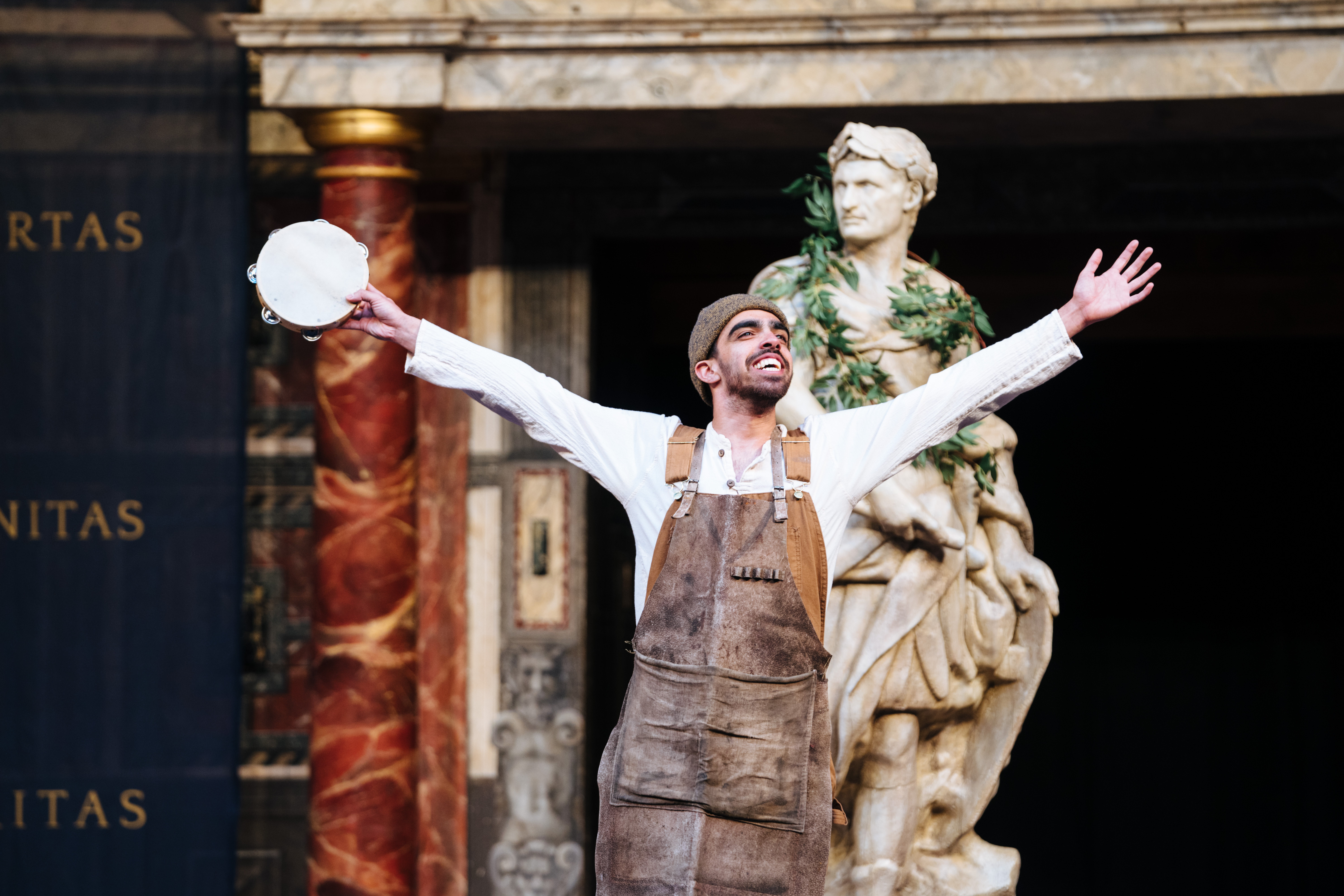 A man in overalls smiles as he puts both of his arms up into the air. Behind him sits a roman statue.