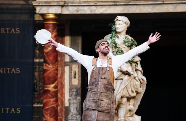 A man in overalls smiles as he puts both of his arms up into the air. Behind him sits a roman statue.
