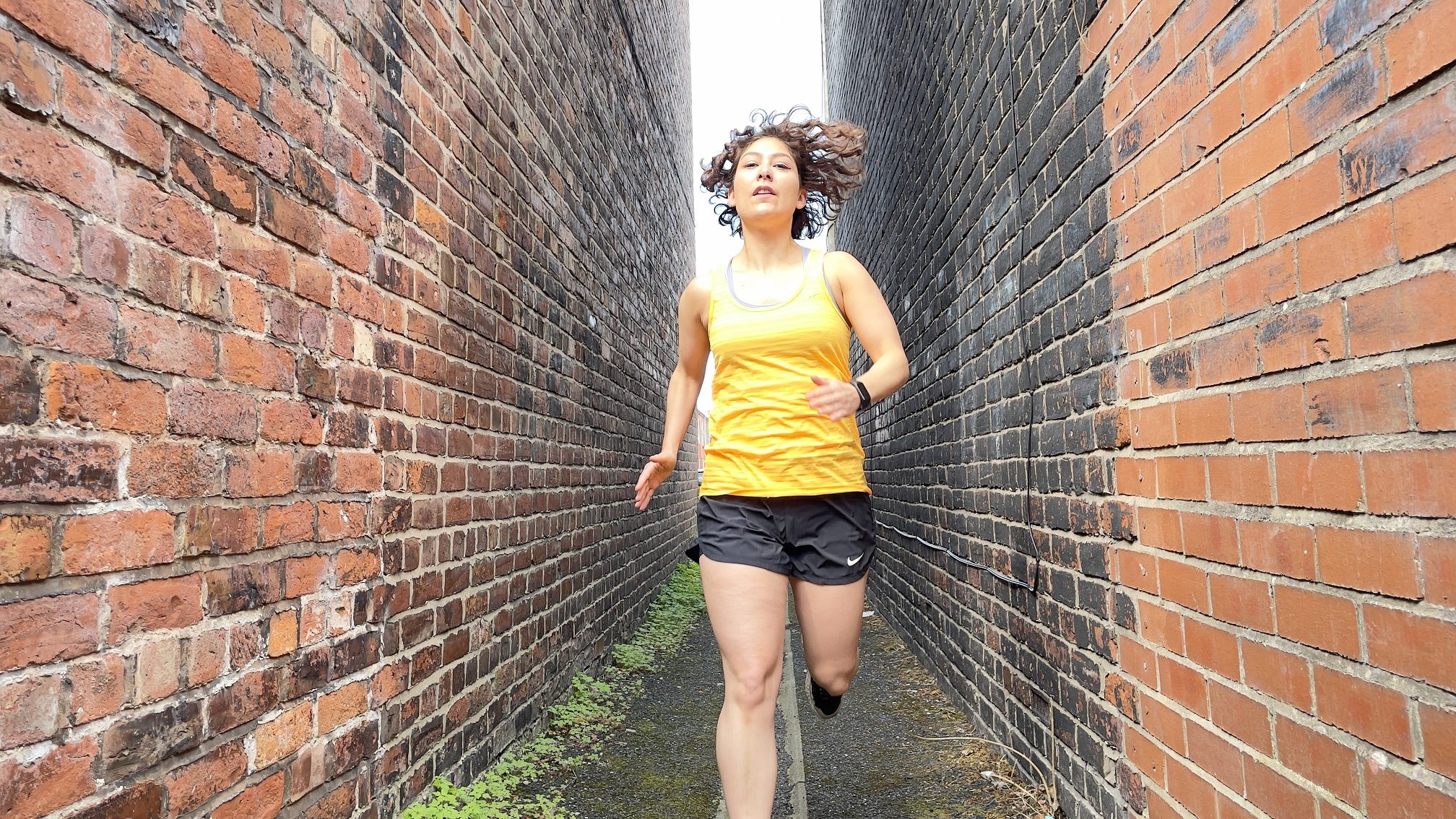 The performer Jenni Jackson running down small alley way, in between two brick walls. Jenni is wearing a running vest and shorts.