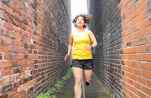 The performer Jenni Jackson running down small alley way, in between two brick walls. Jenni is wearing a running vest and shorts.