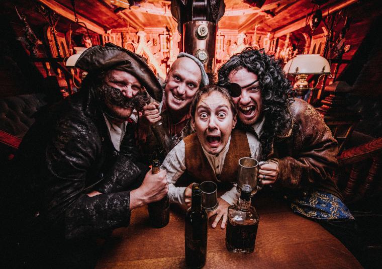 4 men dressed as pirates are set on a wooden table with whisky bottles. They are all pulling exaggerated expressions towards the camera.