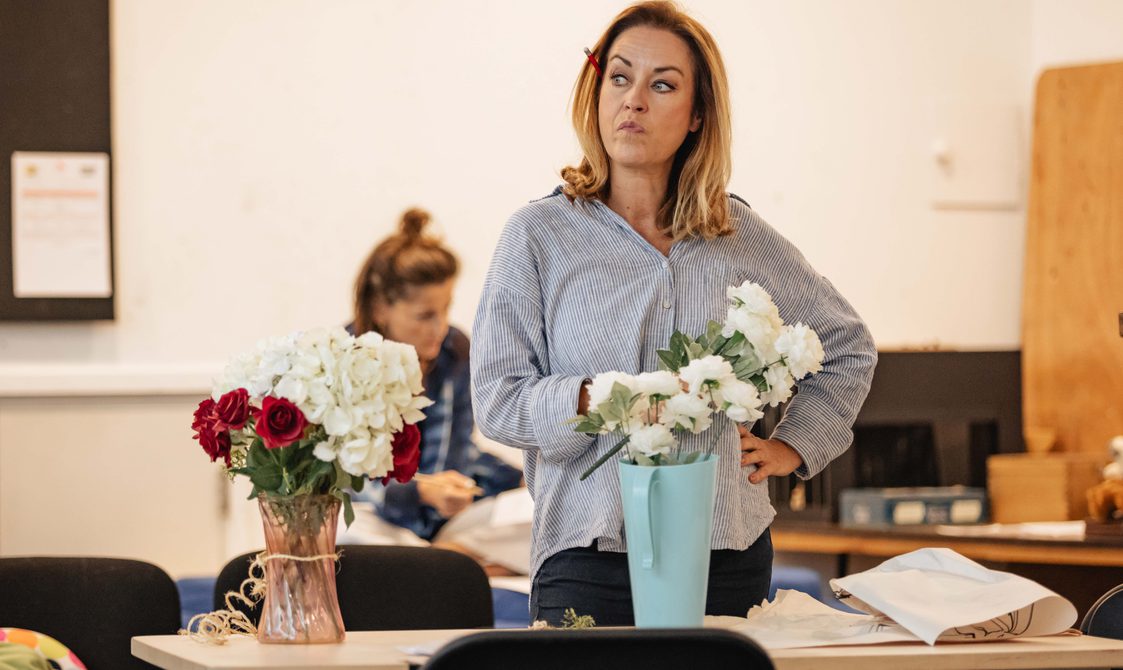 A woman with blonde hair wearing a blue shirt stands behind a table of flowers, she looks off to the side