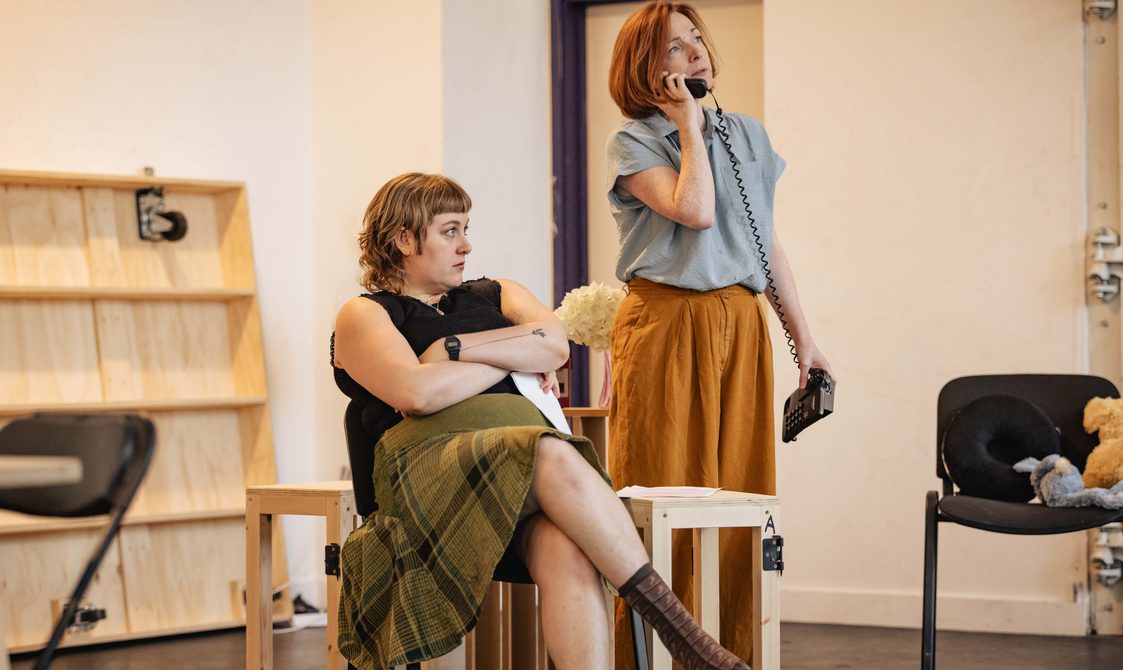 A woman sits crossed legged on a chair, another woman holds a wired phone up to her ear.