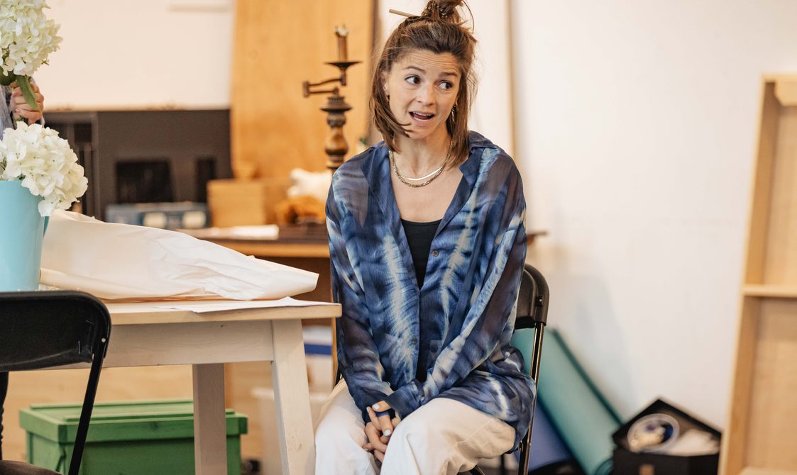 A woman with brown hair and a blue shirt sits in a chair, she has her mouth open and looks off to the side.
