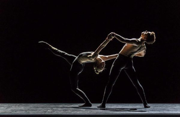 Two ballet dancers on stage, one leaning backwards with leg kicked up in the air while being supported by a dancer behind holding their hands.