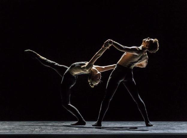 Two ballet dancers on stage, one leaning backwards with leg kicked up in the air while being supported by a dancer behind holding their hands.