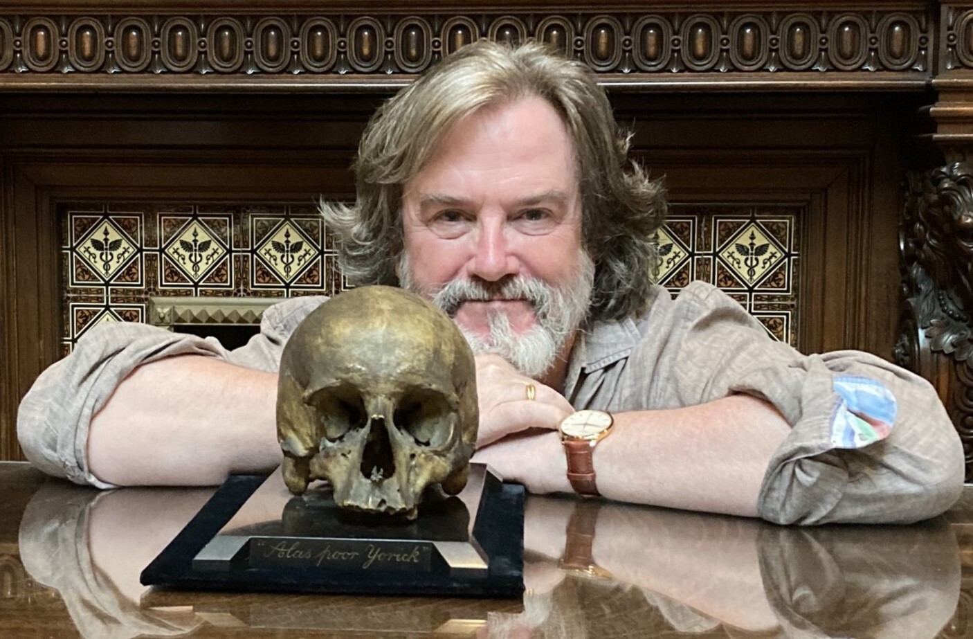 Gregory Doran, a man with medium length brown hair and a grey beard, rests his forearms on a desk in front of him. There is a skull on the desk with the words "Alas, poor Yorick" engraved on the front of the base.