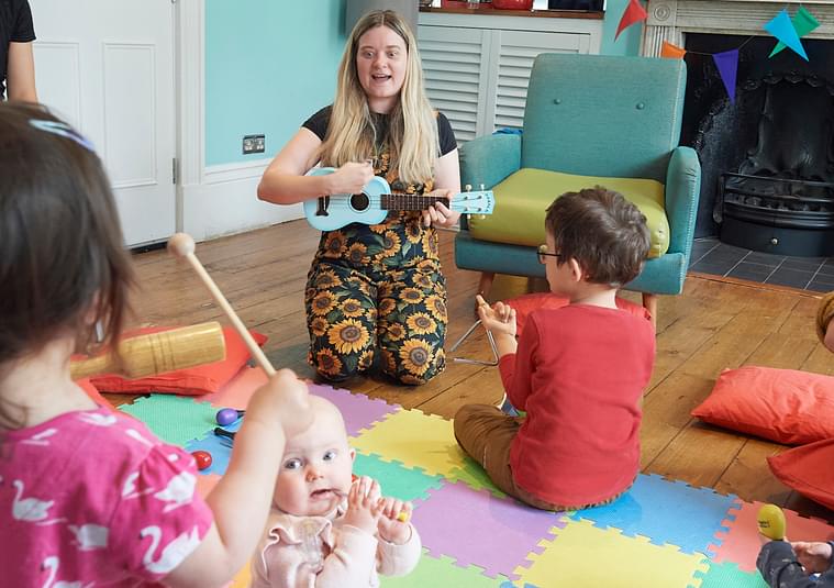 Image is of a Hey Diddle Diddle session, with young babies and toddlers in the middle, either standing or sitting on colourful floor matting, with parents sat around them. A woman is knelt down singing and playing guitar to the children.