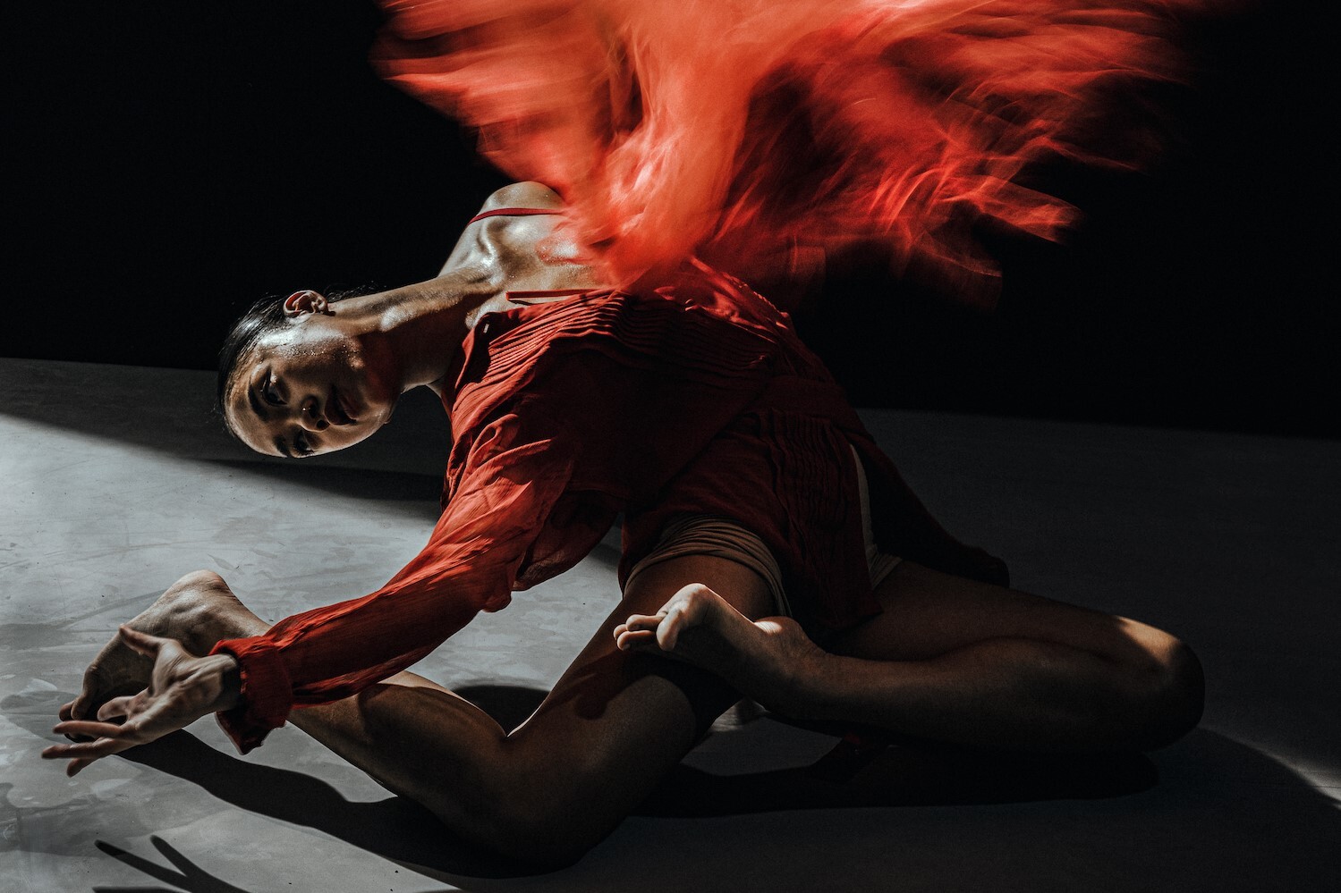 A woman wearing a red dress is on the floor arching backwards with one arm out reached and her head leaning back. Red smoke is coming off of her body