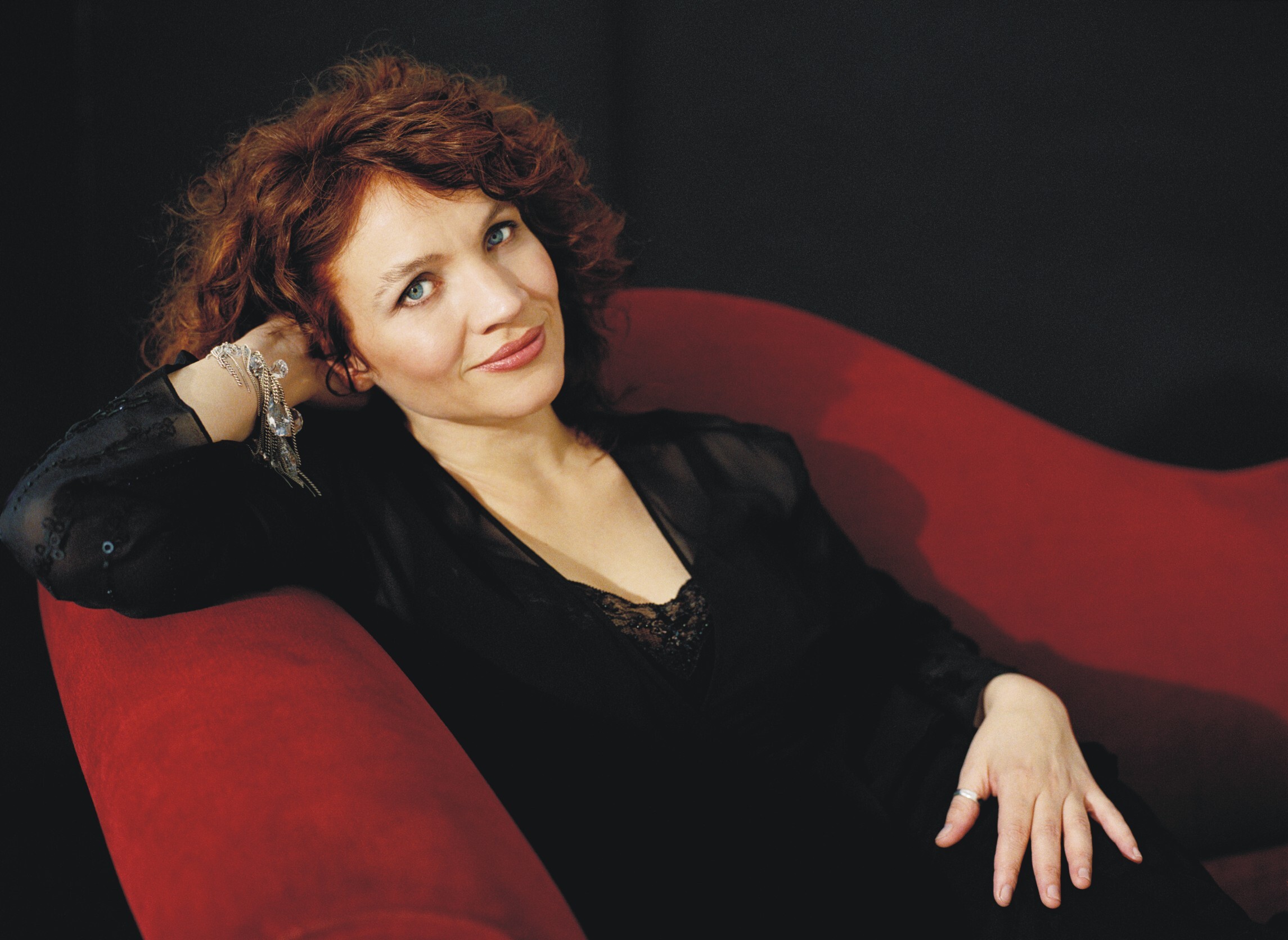 An image of Jacqui Dankworth lying on a red chair with one hand behind her head. She is wearing a black dress.