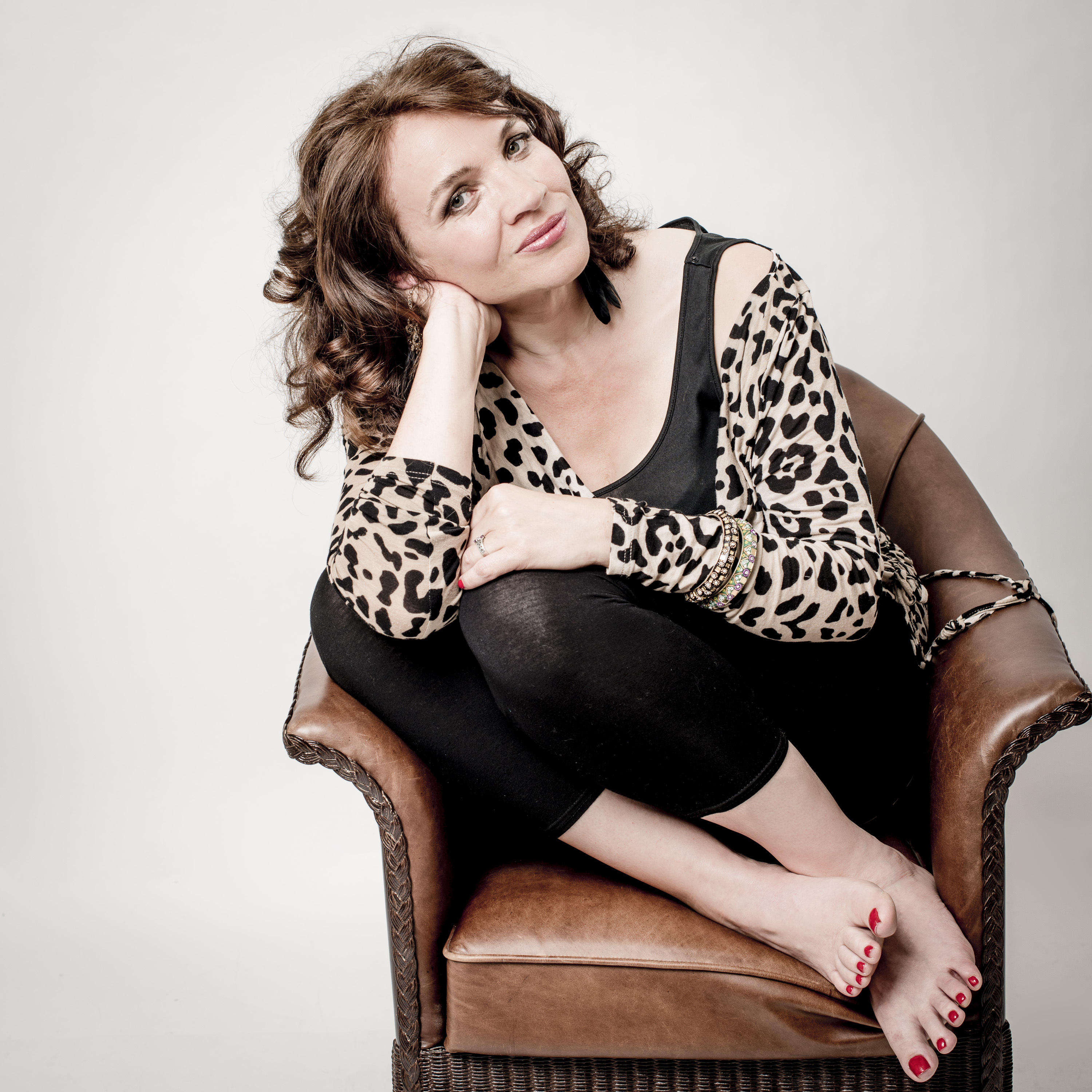 Image of Jacqui Dankworth sat with her legs tucked up on a brown leather chair with her head titled resting on her hand. She looks towards the camera. She is wearing a leopard print cardigan and black top. Her hair is shoulder length and curly.