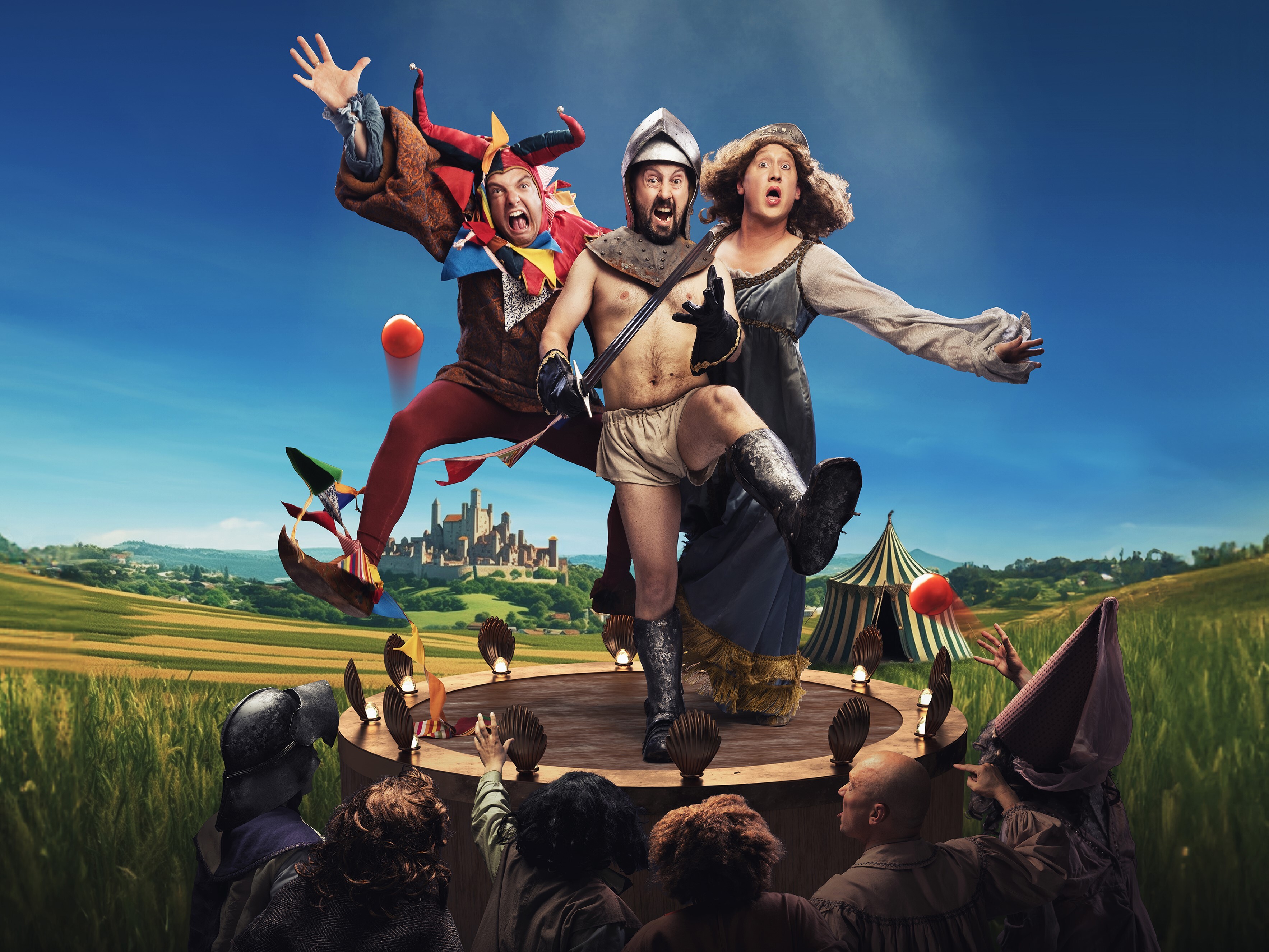 A jester, a knight in underwear and a maiden are standing on top of a wooden circular structure. There are six people below them looking up at them. A medieval tent and view of the town and countryside are in the background.