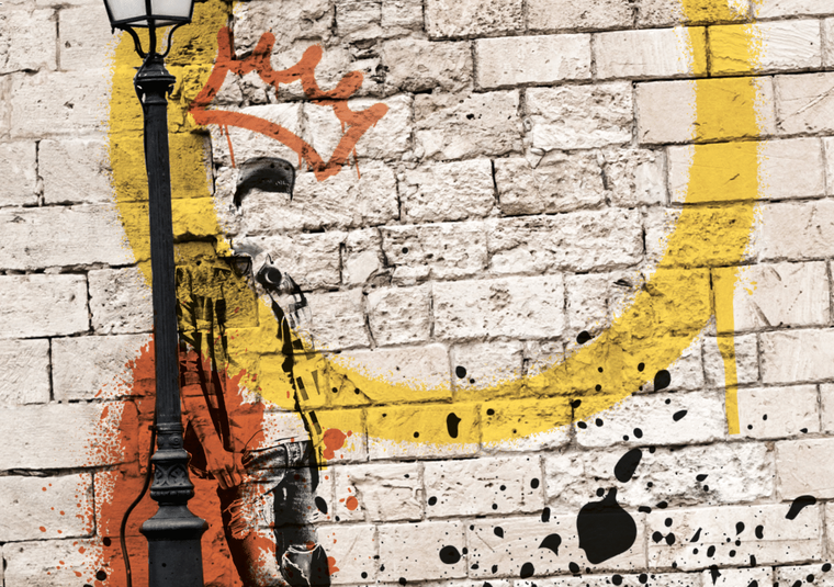 A brick wall spray painted with a yellow circle and orange crown above a graffitied silhouette of a man. He leans against a lamppost.