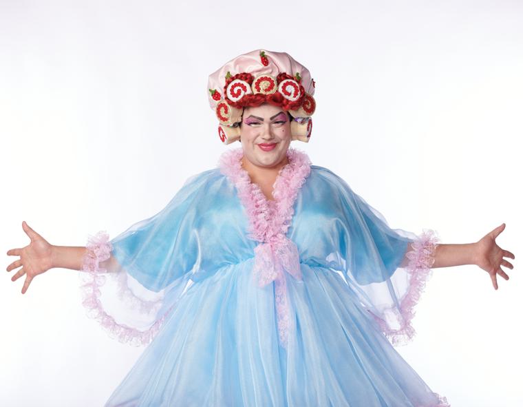 Alasdair Buchan dressed as a Dame. He wears a blue tulle night dress with a fluffy pink collar. He wears a silky looking head dress on with swiss roles round the edge.
