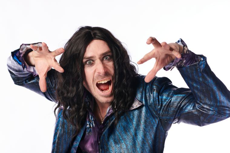 Robin Hemmings in a long black wig with his mouth open and hands clawed to either side of his face.