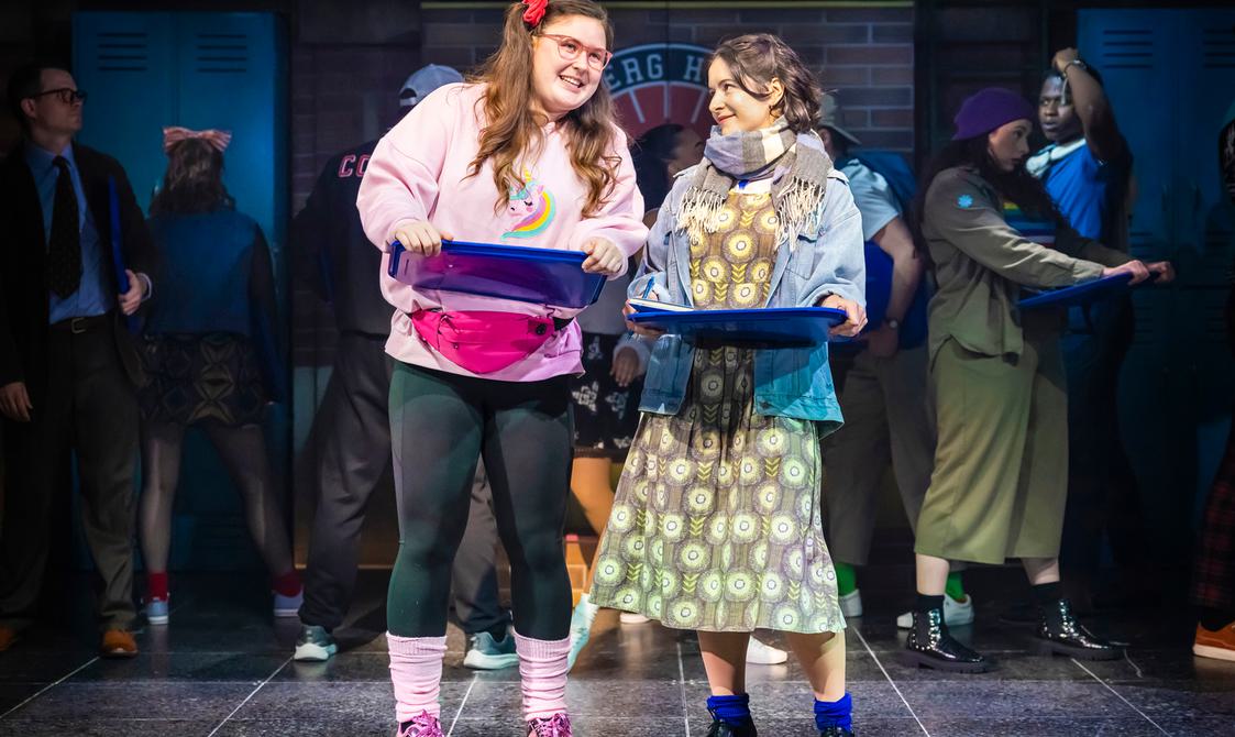 Two women stood together holding dark blue trays, one wearing pink rolled down socks and sparkly trainers with black leggings and a sweatshirt. Other woman is wearing a yellow/beige/cream patterned dress with a denim blue jacket and stripy scarf around her neck. Royal blue rolled down socks with black small heeled, laced up shoes.