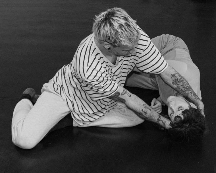 A black and white image of two dancers on the floor, one kneels looking down at another and supporting their head as they lay down on the floor.