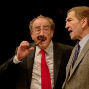 Two men on stage. One has a cigar in his mouth.