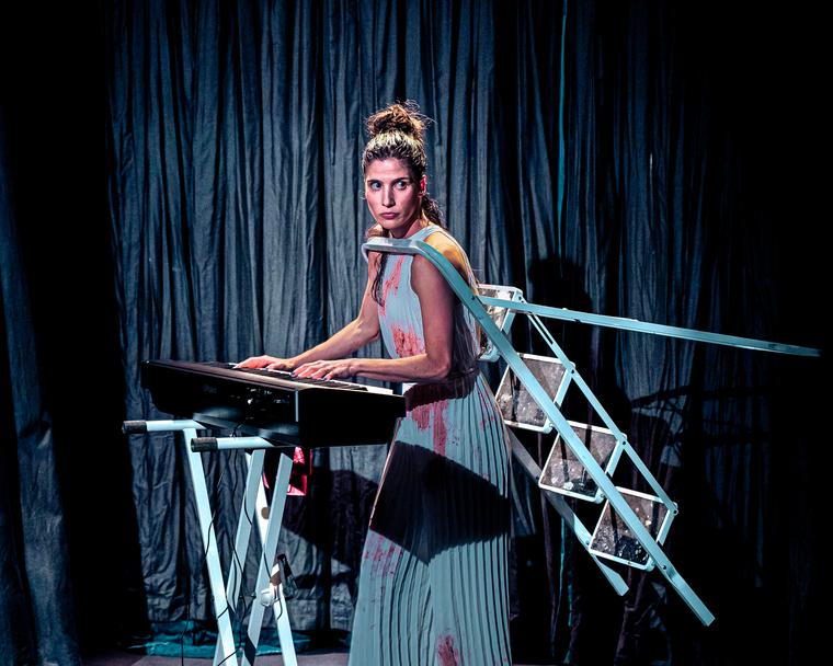 A woman with a brown bun stands in front of a keyboard with a light blue step ladder hooked over her shoulders.