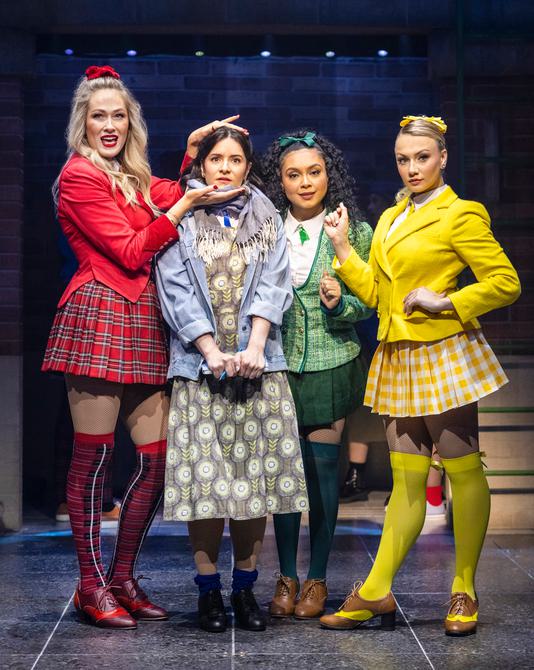 Four women stood together. The left person is wearing a red ribbon in her long blonde hair. A red jacket cut short to the waist with pointed edges at the front, a red check mini skirt, check knee highs and red and maroon laced up heeled shoes. The second person is wearing a dress with a denim blue jacket and stripey scarf around her neck. Royal blue rolled down socks with black small heeled, laced up shoes. The third person is wearing a green ribbon in her long dark brown hair. A white shirt with a green small tie at the collar and a green checked jacket with silver buttons on. A green mini skirt and green knee highs with brown laced up shoes.The fourth person has her blonde hair tied up with a yellow ribbon. A white shirt with a yellow small tie at the collar and a plain yellow short jacket. A yellow and white pleated mini skirt, bright yellow knee highs with brown and yellow laced up shoes.