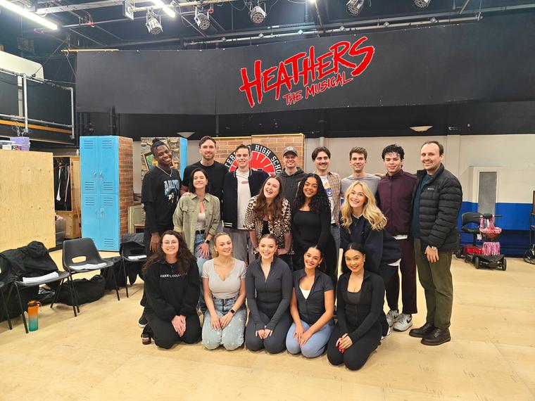 Three rows of people stood in a rehearsal room smiling at the camera. 'Heathers the Musical' is written above them