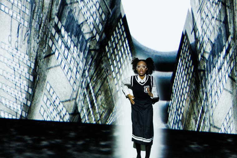 A young girl is running in an old fashioned school uniform, a surreal green grey city of sky scrapers projected behind.