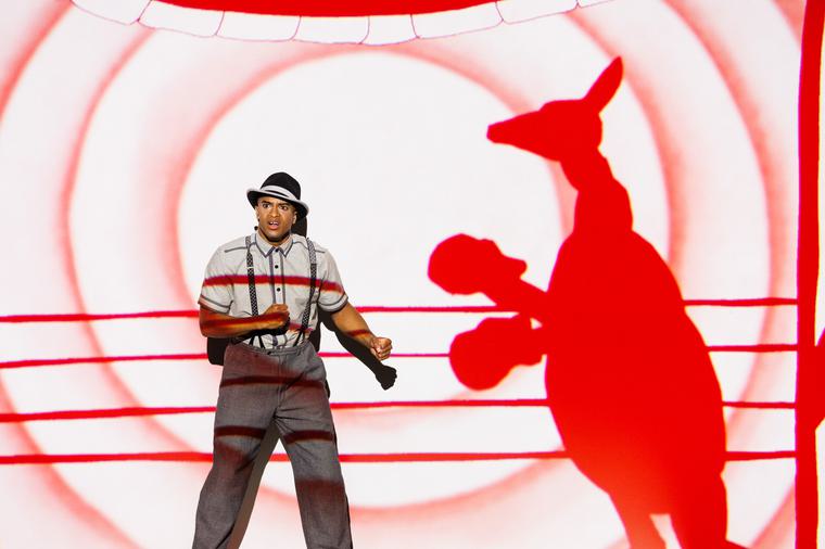 A man dressed in old fashioned clothes (braces and hat), has his hands clenched in a punching position. Next to him is a red projection of a silhouette of a kangaroo wearing boxing gloves with the lines of the boxing ring projecting across.