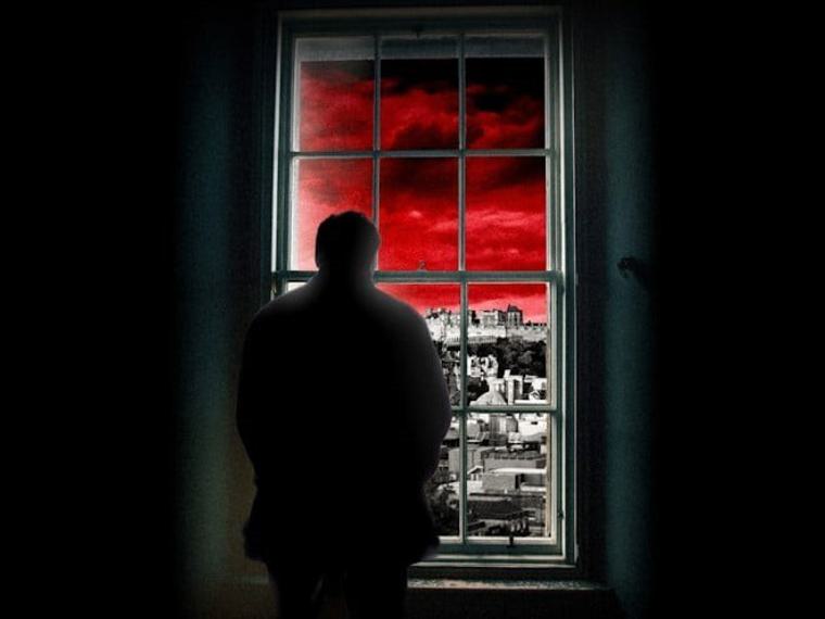 A man is stood looking out of a window, inside everything is dark and grey, outside is a black and white city scape under a bright red stormy sky.