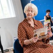 Susie Blake looks up from a magazine. Other members of the cast stand behind her in the distance.