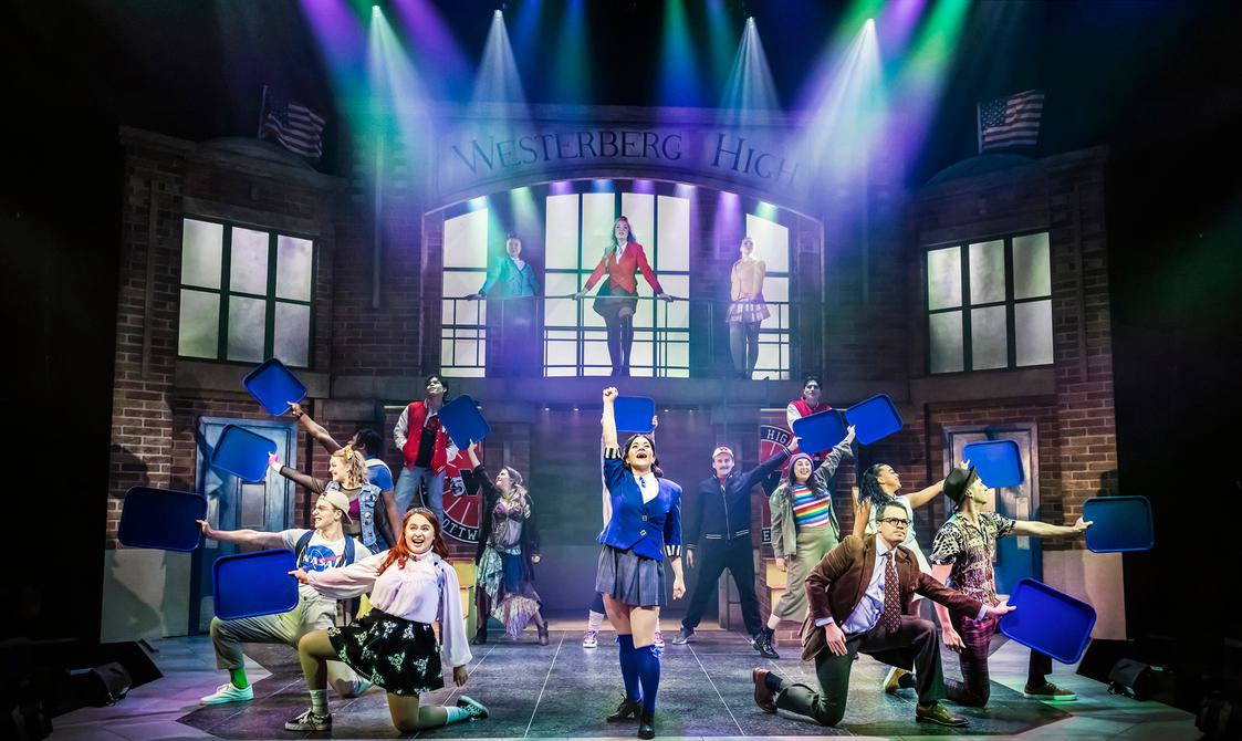 Full cast on stage with one woman stood up holding up her fisted right arm. She is in the middle of the rest of the cast who are kneeling on one leg who are all holding up high a blue tray. Westerberg High is lit up high in the background with three woman stood underneath it.