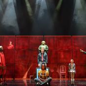 A judge stands behind two people. Two people dressed as barristers stand either side. A woman sits on a chair at the back of the stage.