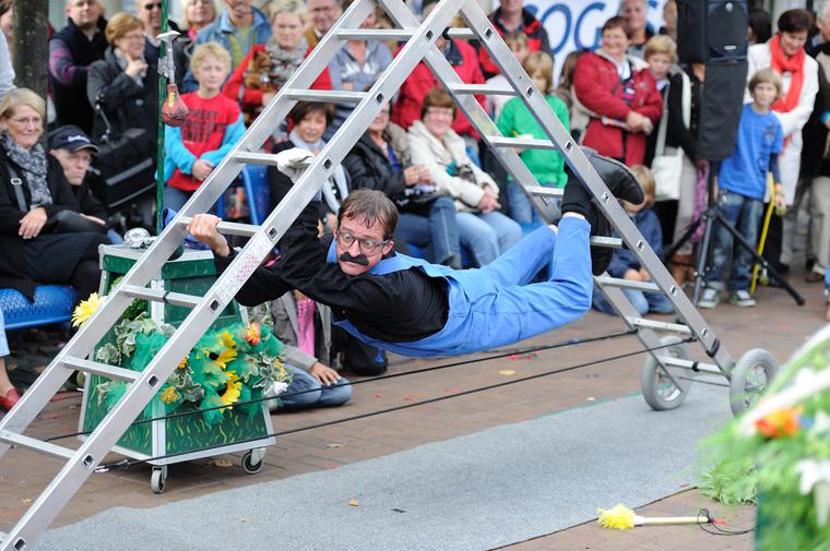 A man in blue dungarees hangs underneath a ladder. Groups of people watch on from behind.