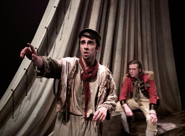 A man dressed in tattered clothing and flat cap raises his arm and looks off to the side of the frame. A man dressed as a soldier sits behind him on a ledge. There is a large sheet behind them in the style of a ship's sail.