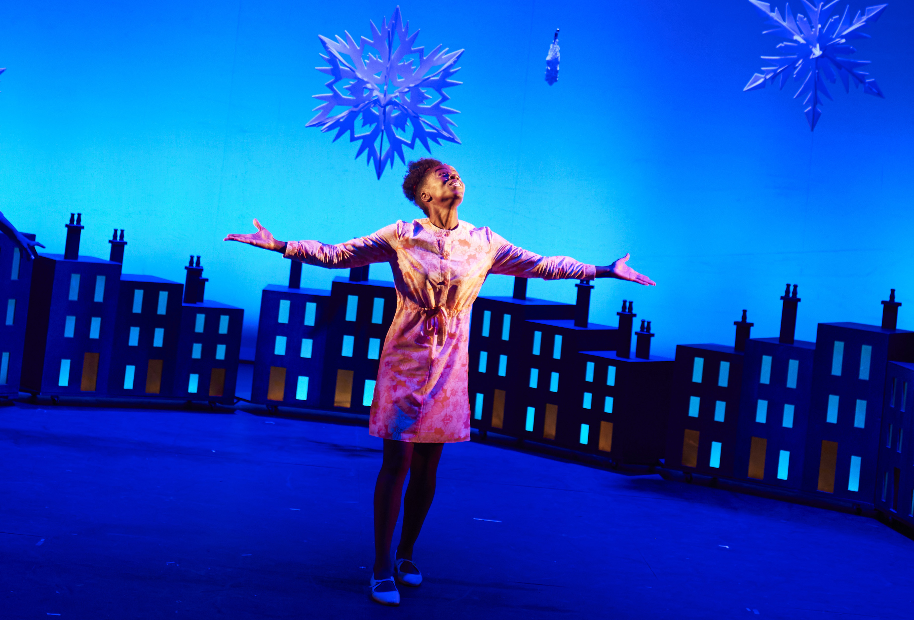 A woman is standing centre stage looking up towards the sky with her arms outstretched. She has her eyes closed and is smiling. In the background is an abstract city scape and in the sky are large icicles.