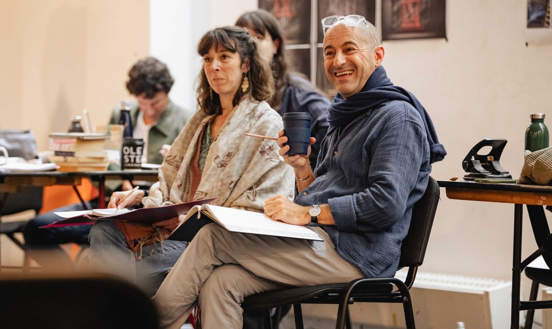 Two people sit in chairs, smiling. They hold scripts on their laps.