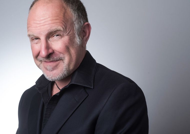 Profile photo of comedian Simon Evans in a dark suit against a grey backdrop.