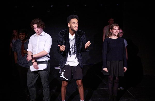 The group of actors stand on stage with the front three in the spotlight. The actor in the front is talking to the crowd energetically.