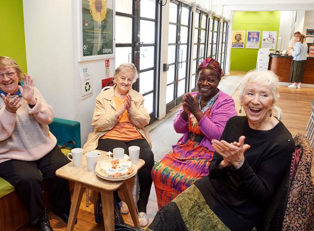 Image shows four woman sat in the theatre foyer around a table with plates and mugs on it. They are all smiling and clapping.