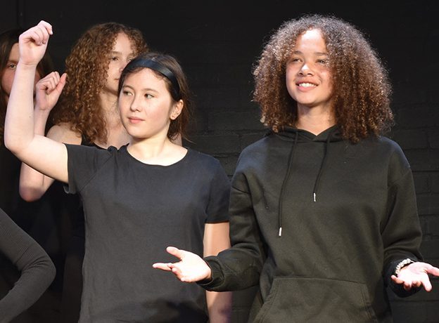 An image of two young people with others in the background. One of them has their hand raised and is looking off to the side and the other is smiling, looking forward with their arms outreached and palms facing upwards.