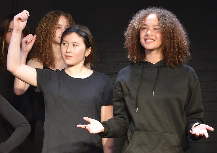 An image of two young people with others in the background. One of them has their hand raised and is looking off to the side and the other is smiling, looking forward with their arms outreached and palms facing upwards.