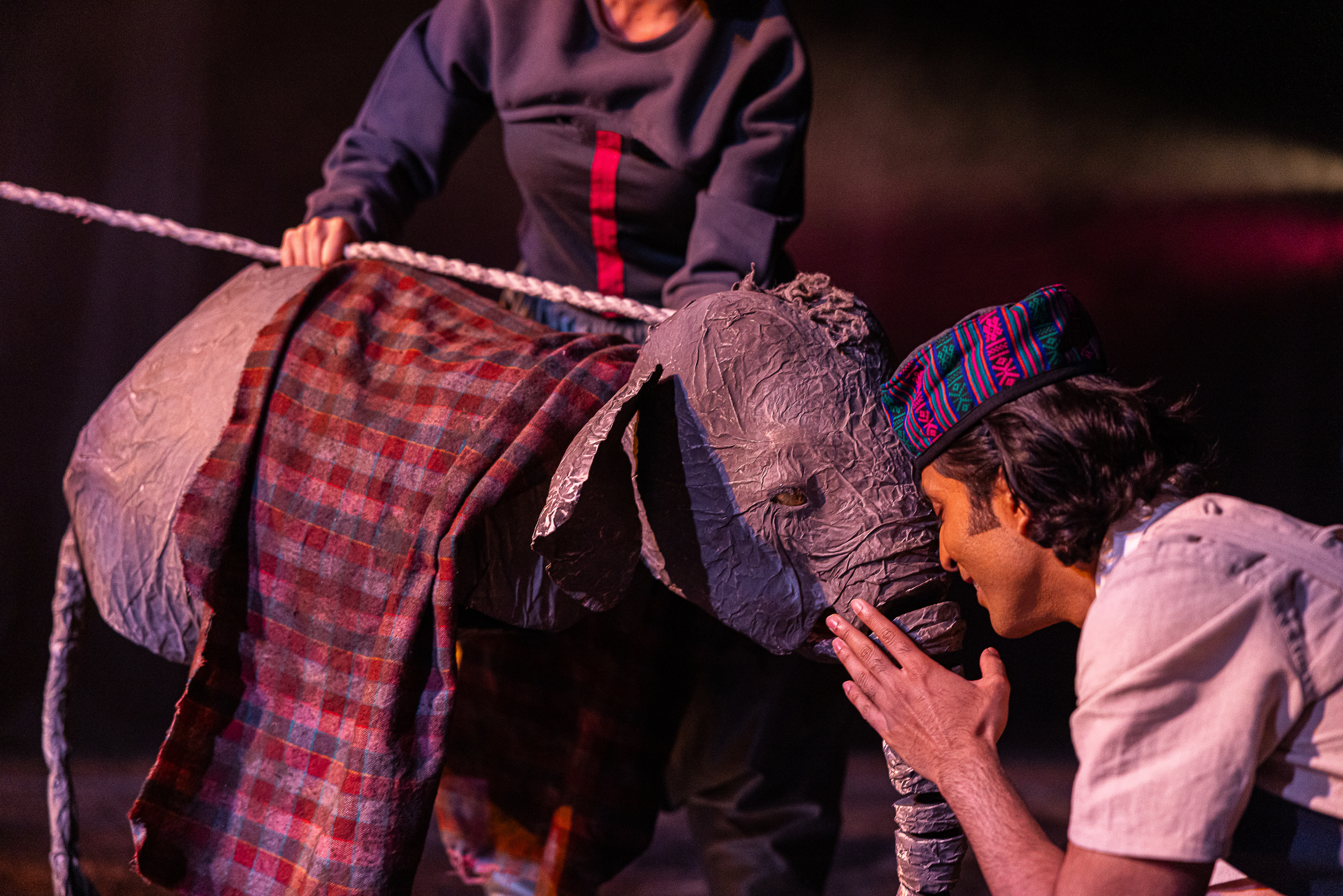 A man holds his face close to the face of a small elephant puppet. The puppet is draped with a red patterned fabric.