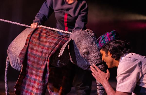 A man holds his face close to the face of a small elephant puppet. The puppet is draped with a red patterned fabric.
